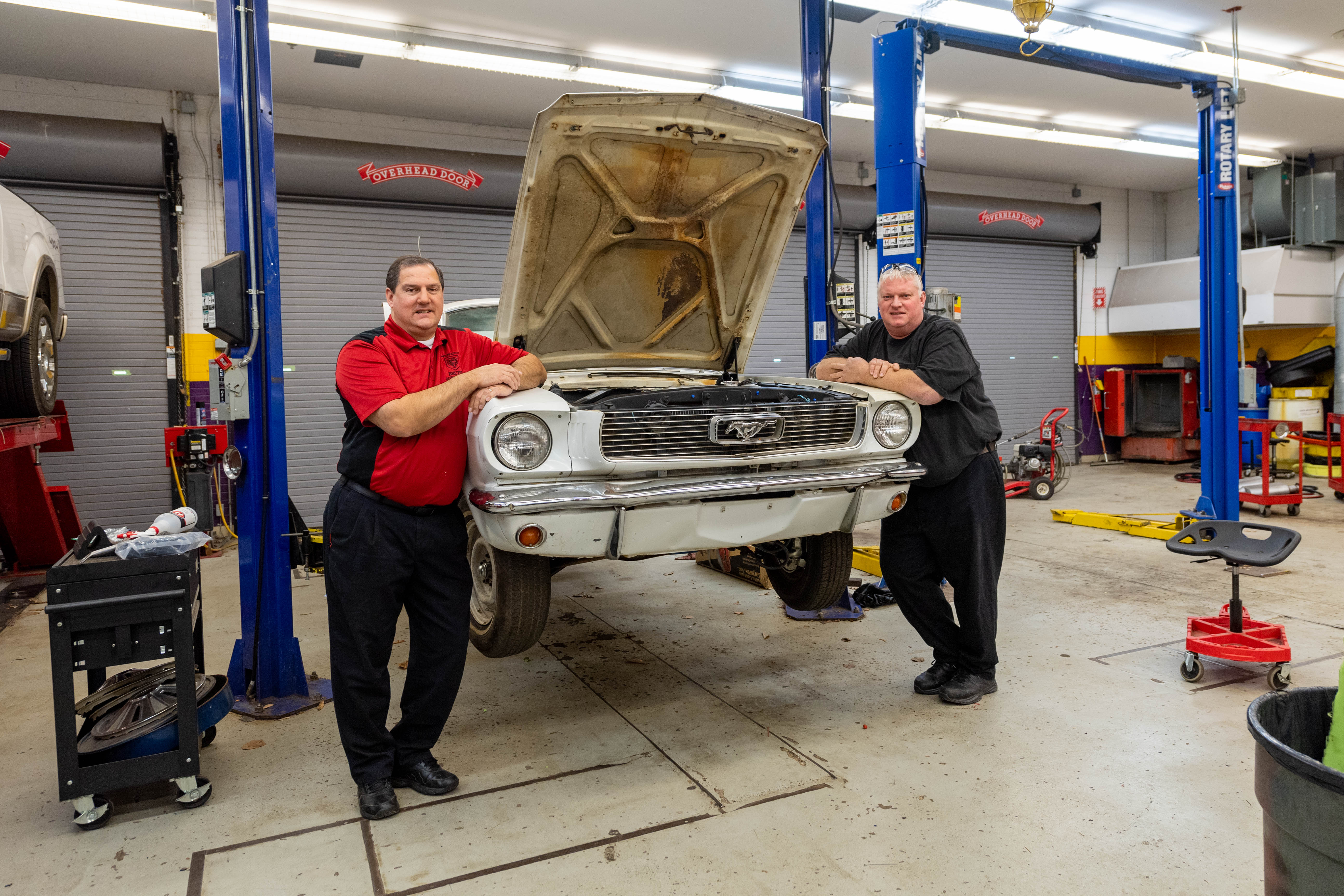 Prakash and Plum stand in front of a Ford Mustang currently being worked on by students in their auto classes.