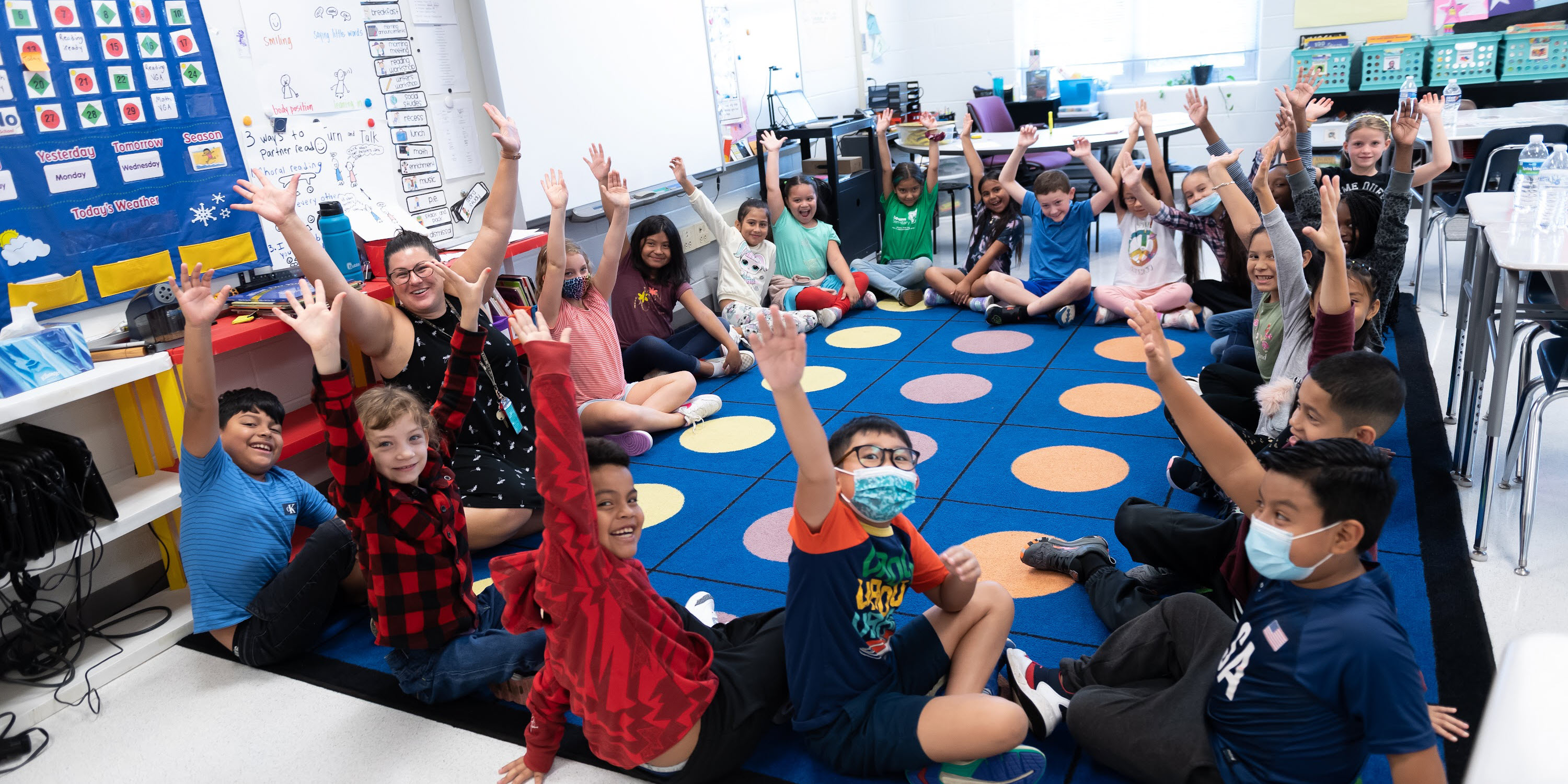 Students sitting on a classroom floor smiling with their hands up