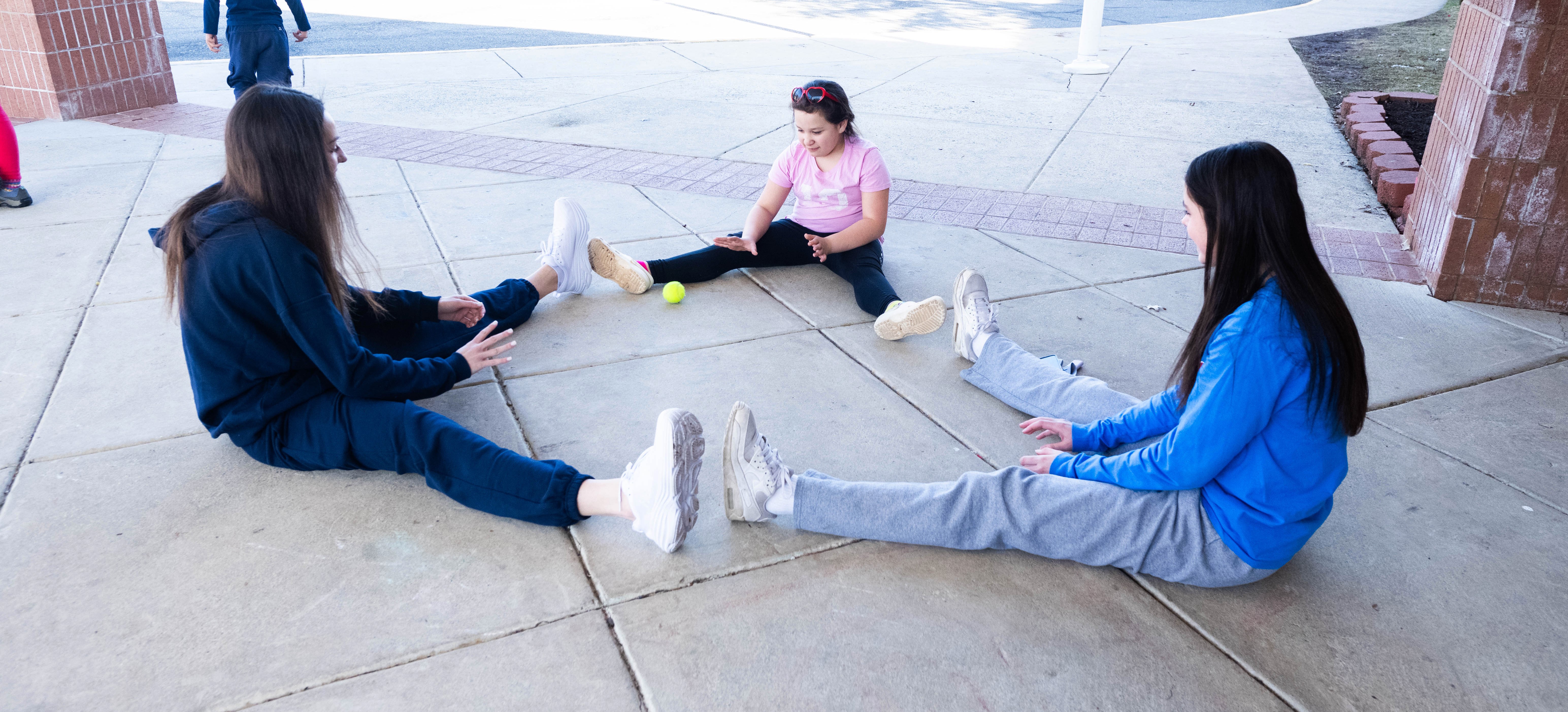 three students sitting on the ground and rolling a tennis ball back and forth