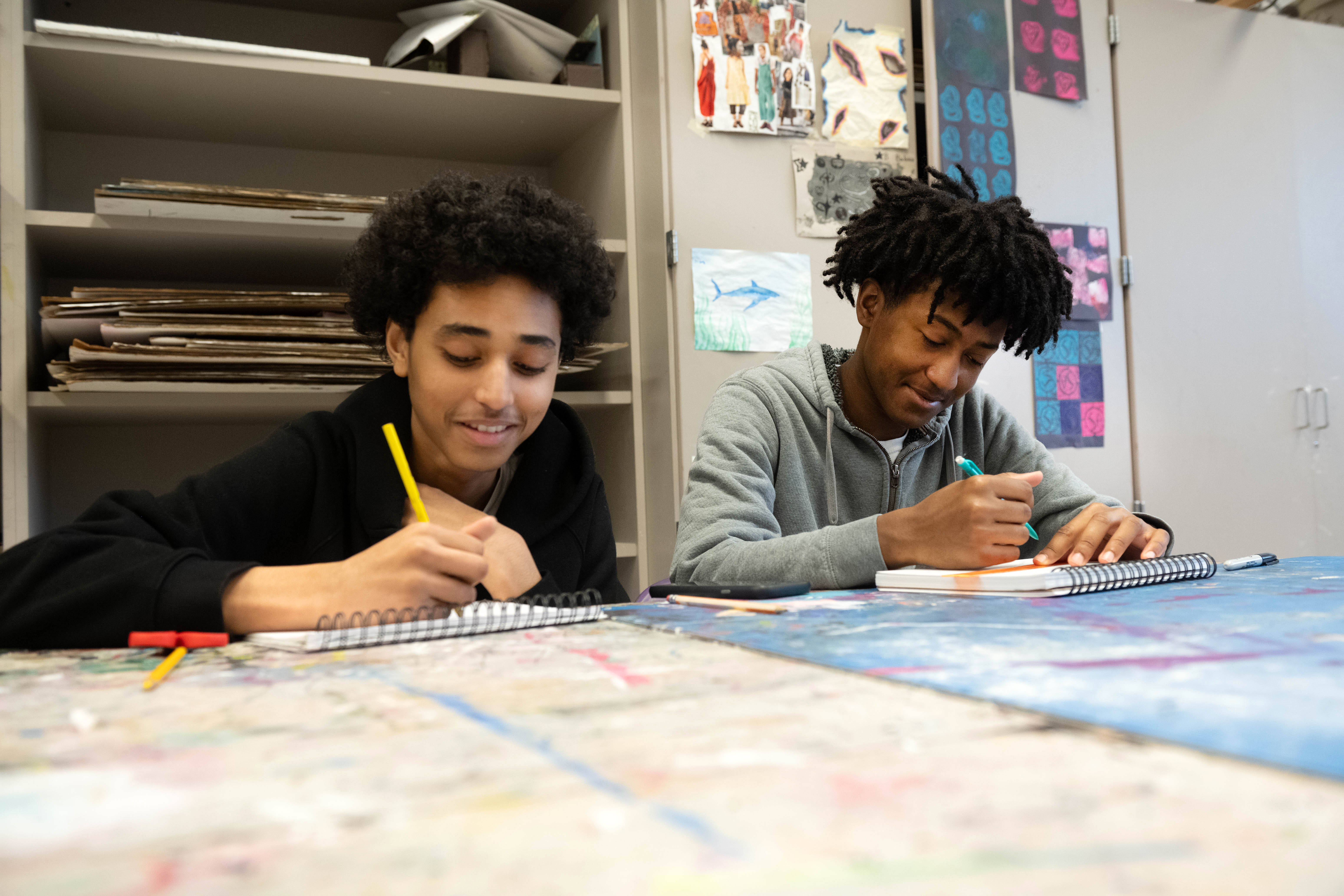 Two students smile as they draw in their sketchbooks.