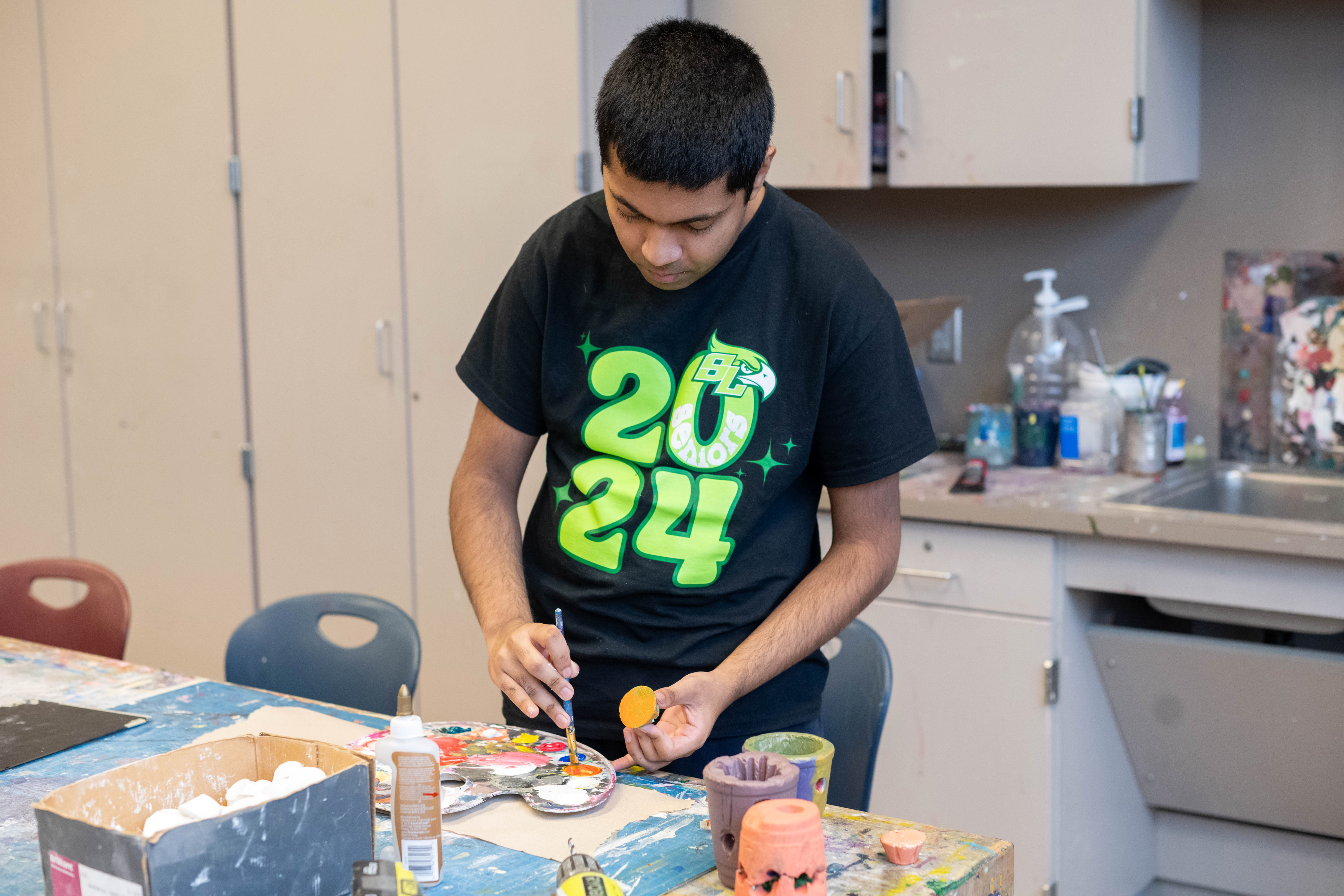 A student paints a small piece of baked clay. he dips his brush into orange paint on a palate. The flat bottom of the clay piece is the same color orange.