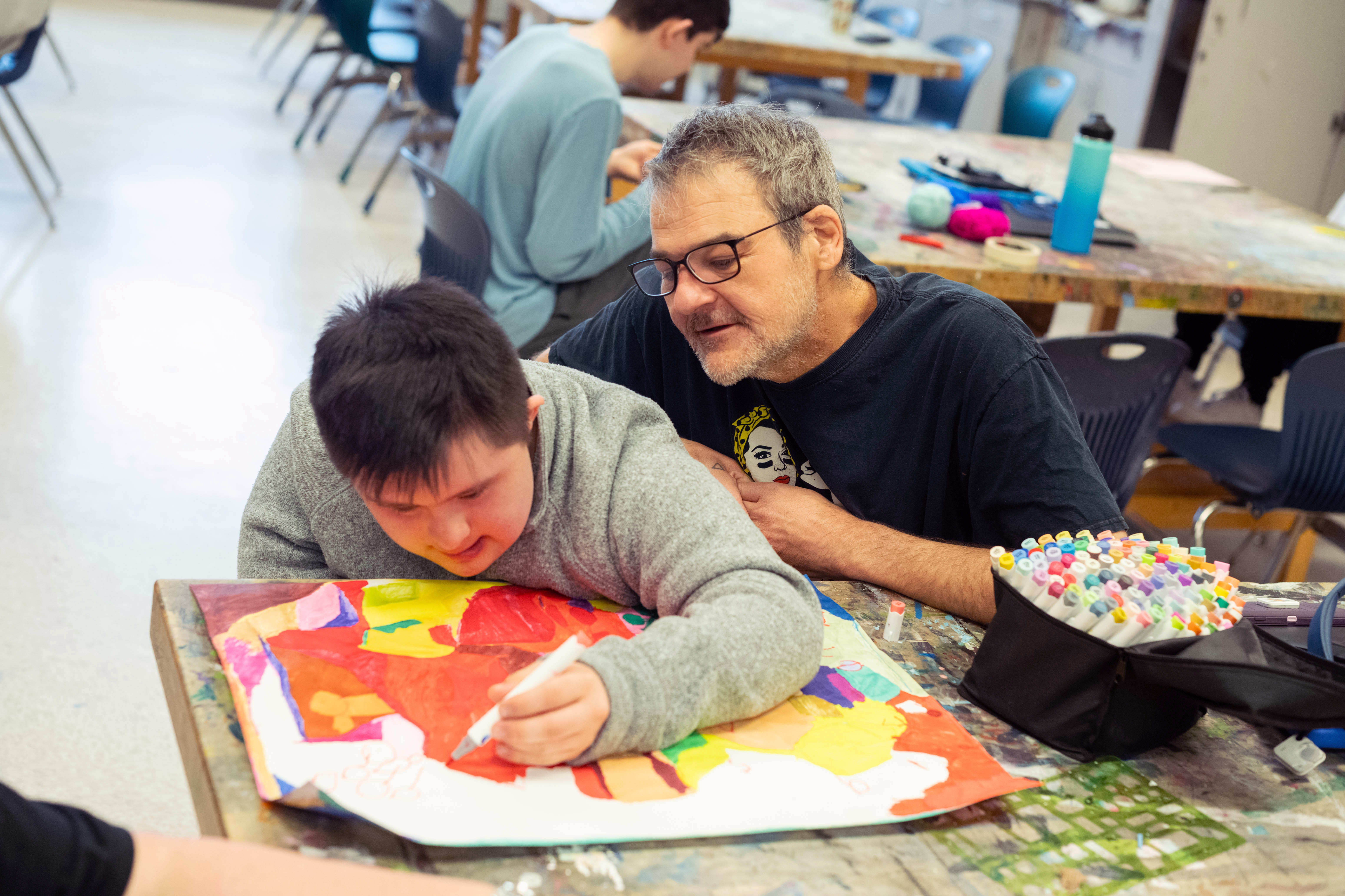 Teacher Matt Ravenstahl looks closely over student Frank Hickok's shoulder as he draws a pattern with colorful markers.