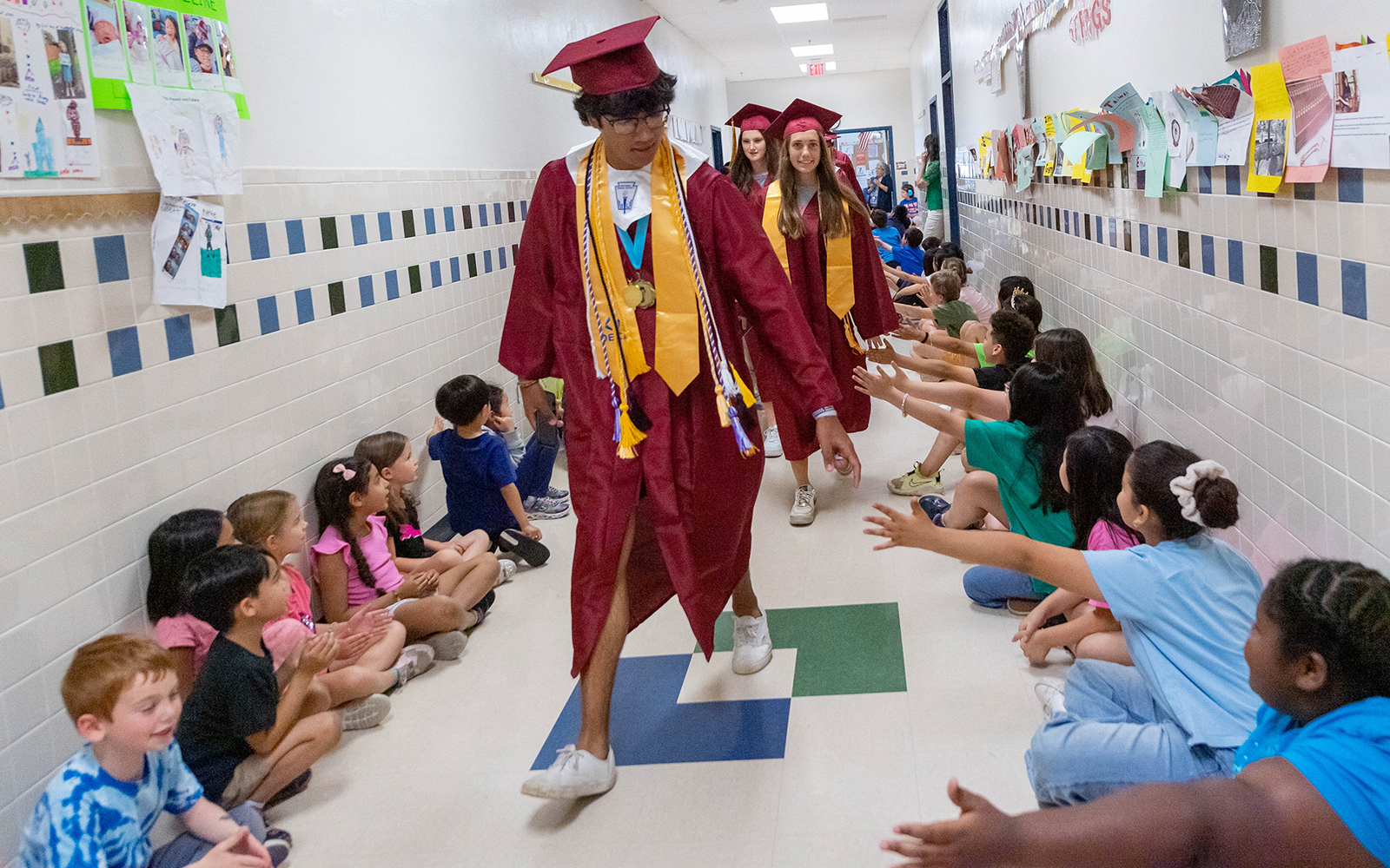 Graduates dressed in gowns walk down the hall of an elementary school