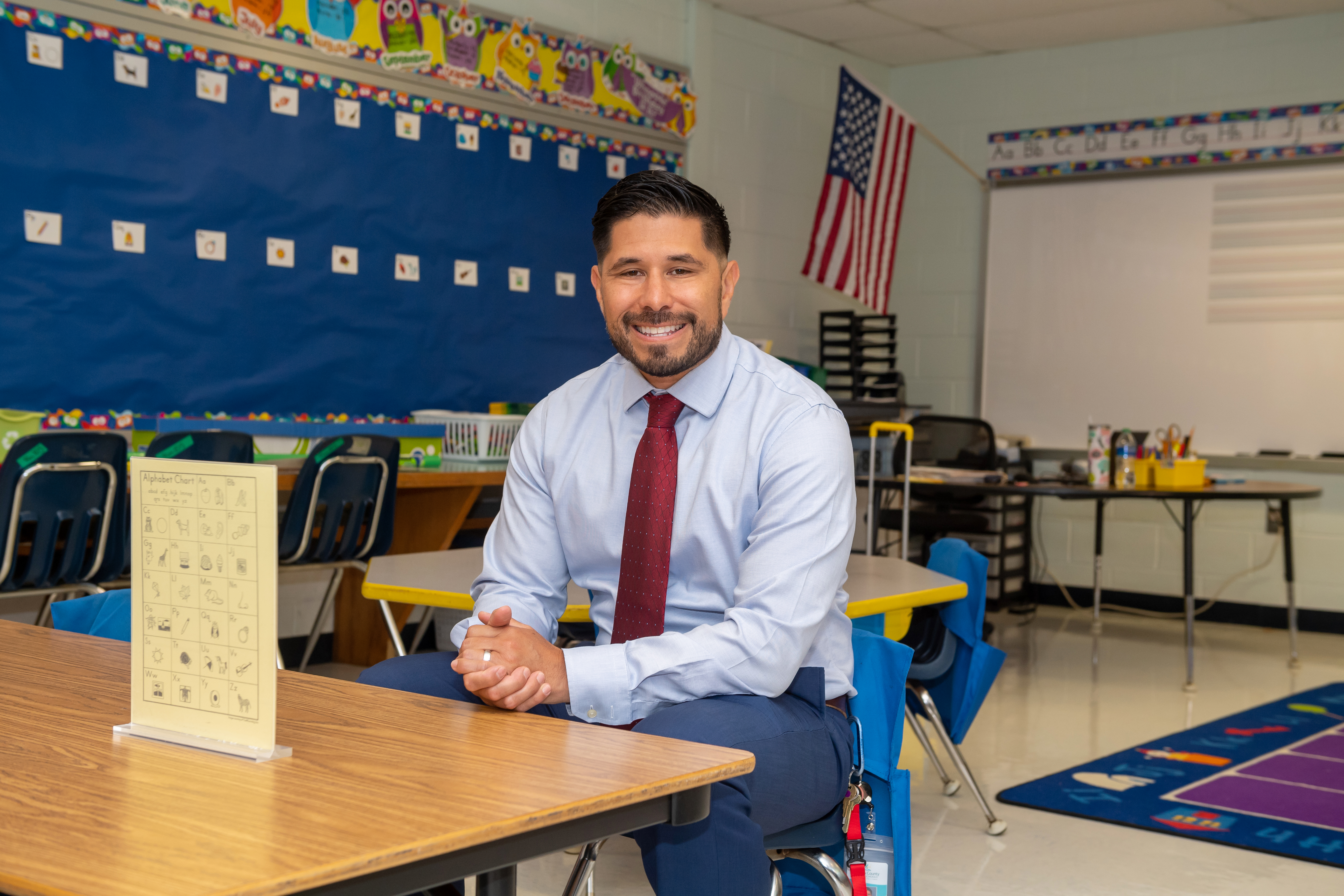 Principal Astudillo sits at a desk in a kindergarten classroom where he was once a student