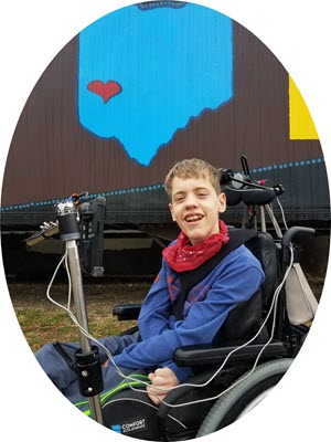 AT Ambassador, Augie, is pictured in a blue shirt with a red bandana sitting in his wheelchair with is communication device