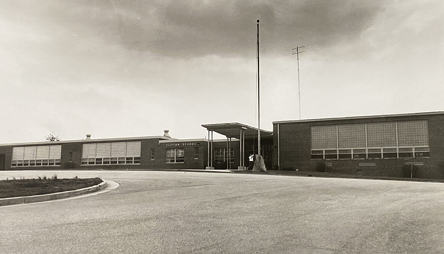 Photograph of the front exterior of Clifton Elementary School taken in the late 1960s. A student is standing by the flagpole.