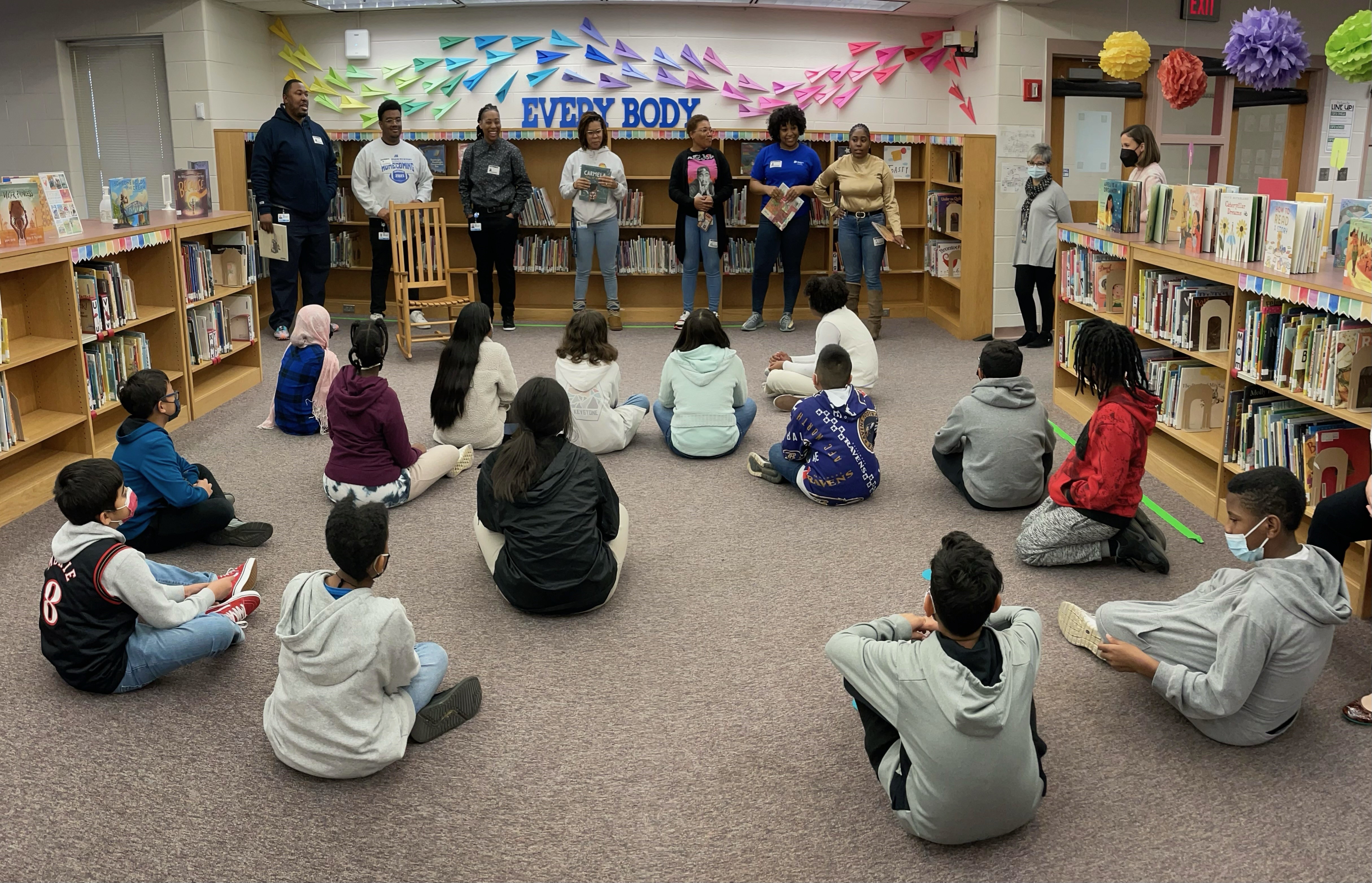 Group of adults reading to a group of students in a school library. 