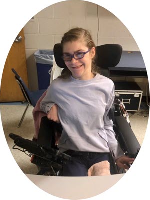 AT Ambassador, Madeline sitting in her wheelchair in a classroom. She is wearing a grey sweatshirt and glasses.