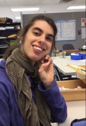 AT Ambassador Graduate, Nicole sitting in her wheelchair in a classroom. She is wearing a blue jacket and gray scarf.