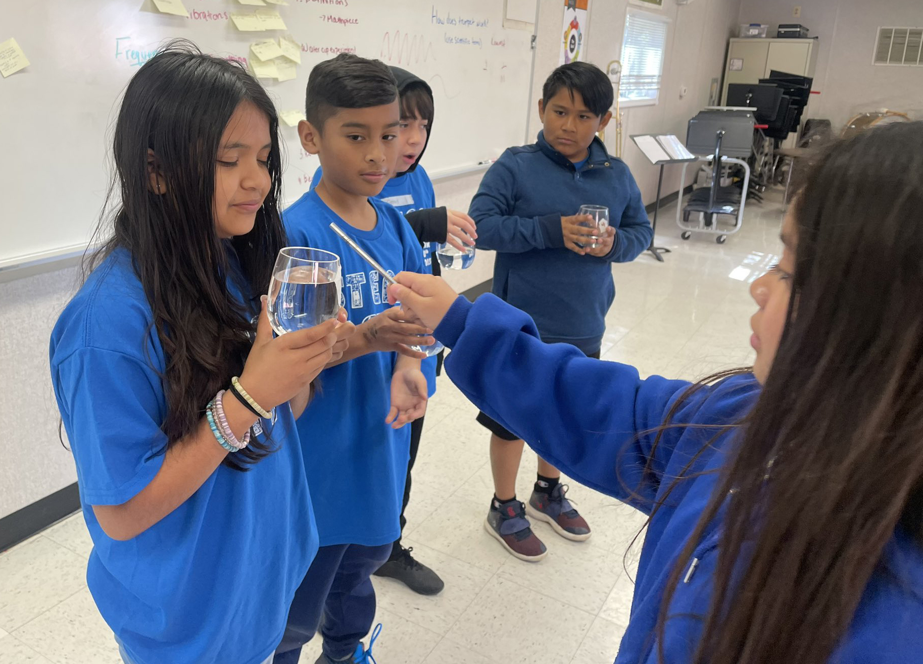 A 6th grade student holds up a glass of water as another student taps a small metal rod at the rim of the glass. Three students stand to watch.
